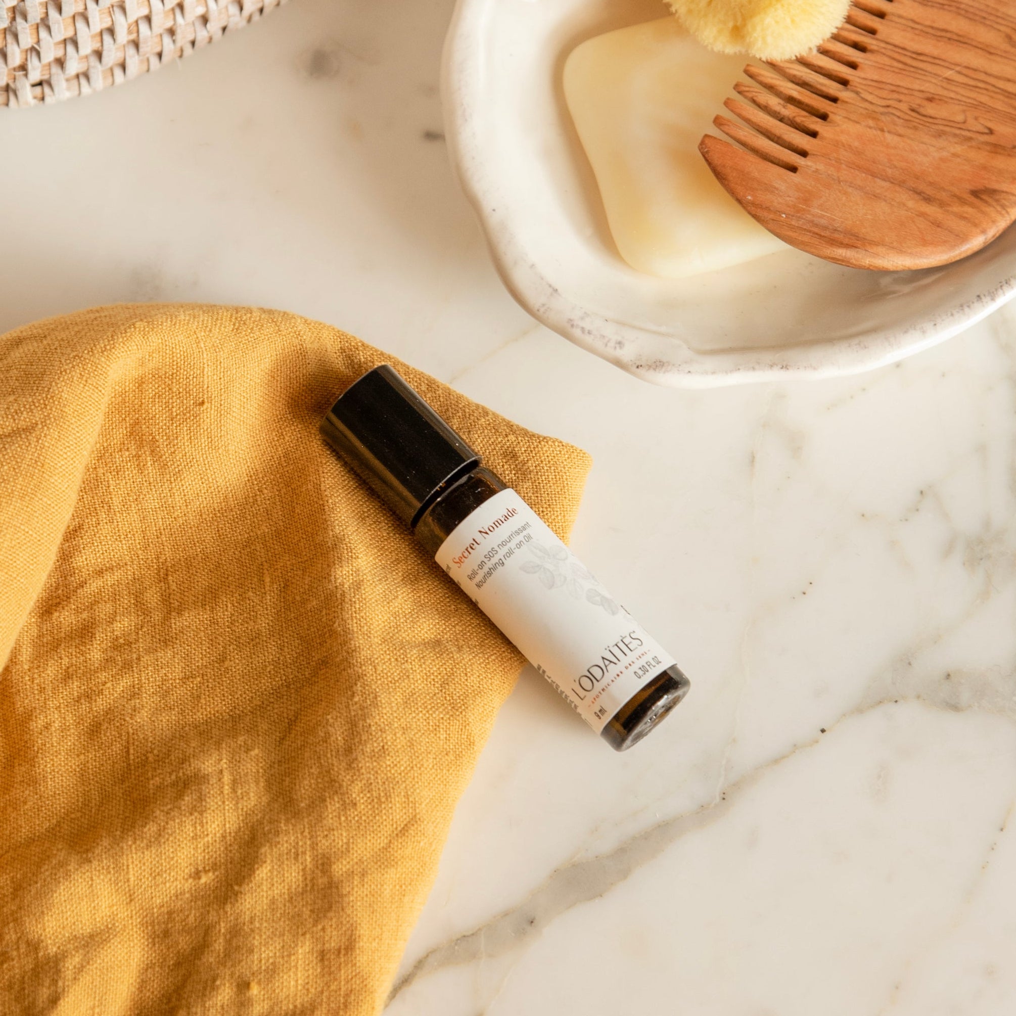 Roll-on SOS nourrissant Secret Nomade de L'Odaïtès sur un torchon jaune à côté d'un porte-savon et d'un peigne