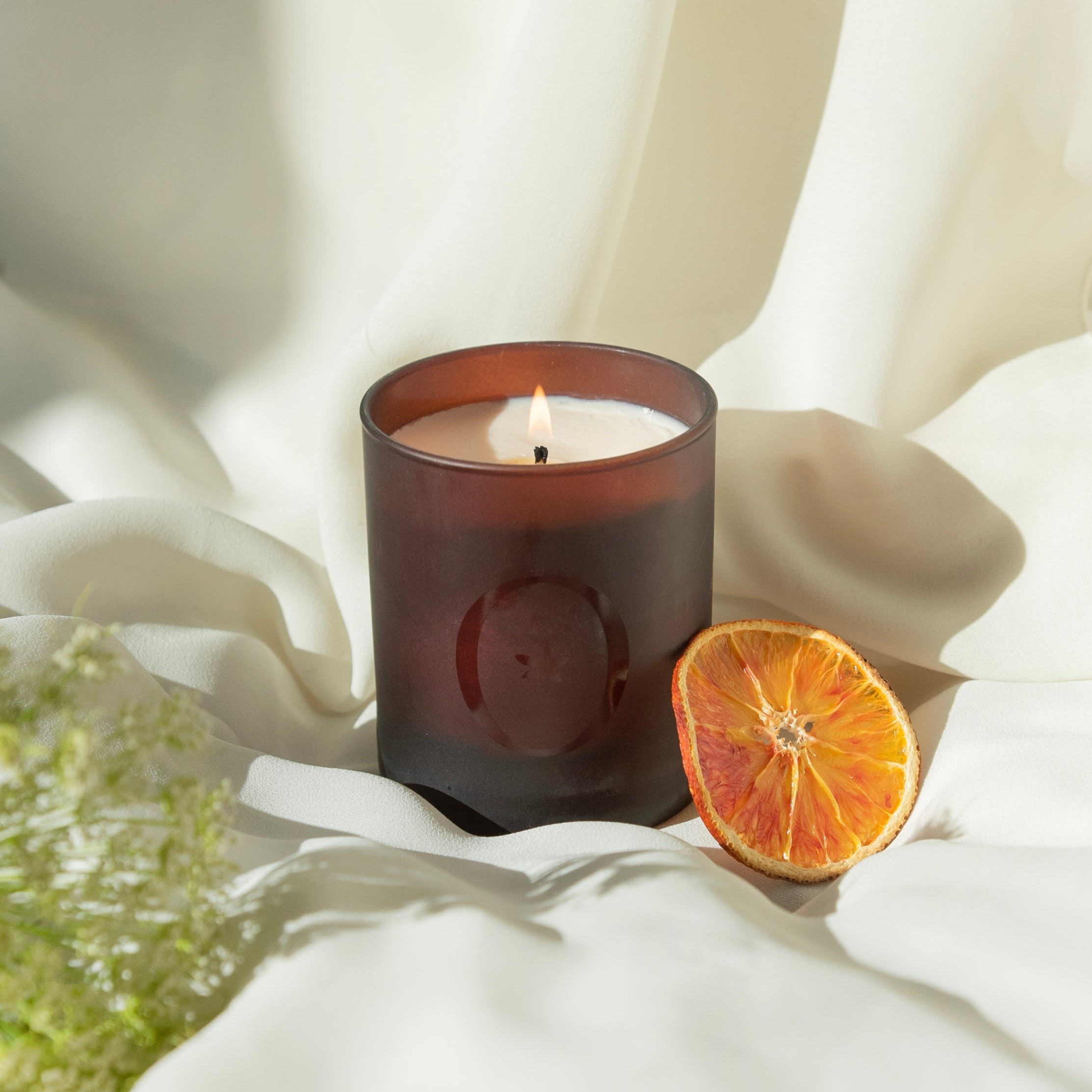Bougie parfumée Orangeraie en Fleur de L'Odaïtès posée sur un drapé avec une rondelle d'orange séchée et une plante verte