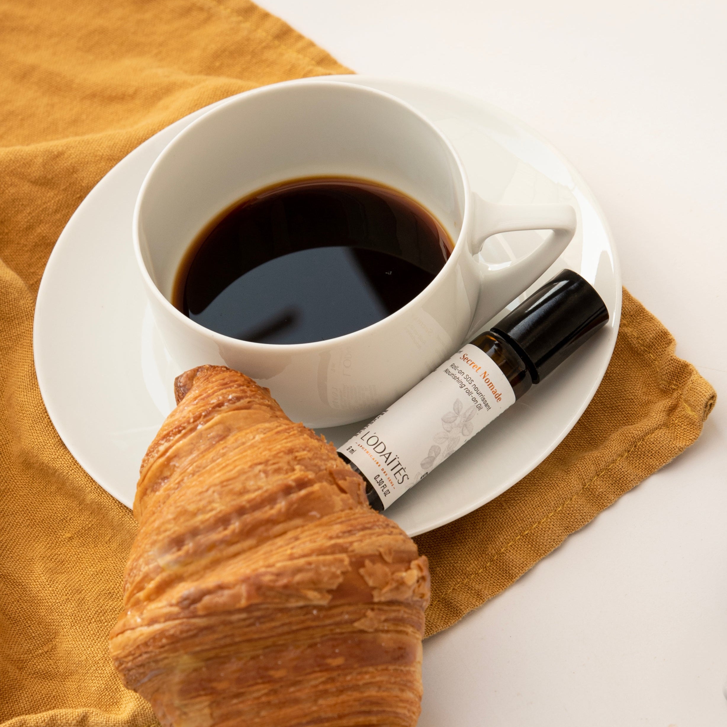 Roll-on SOS nourrissant Secret Nomade de L'Odaïtès posé à côté d'une tasse de café et d'un croissant
